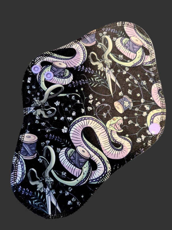 Black background with green snake twined around thread and scissors with branches with tiny white flowers